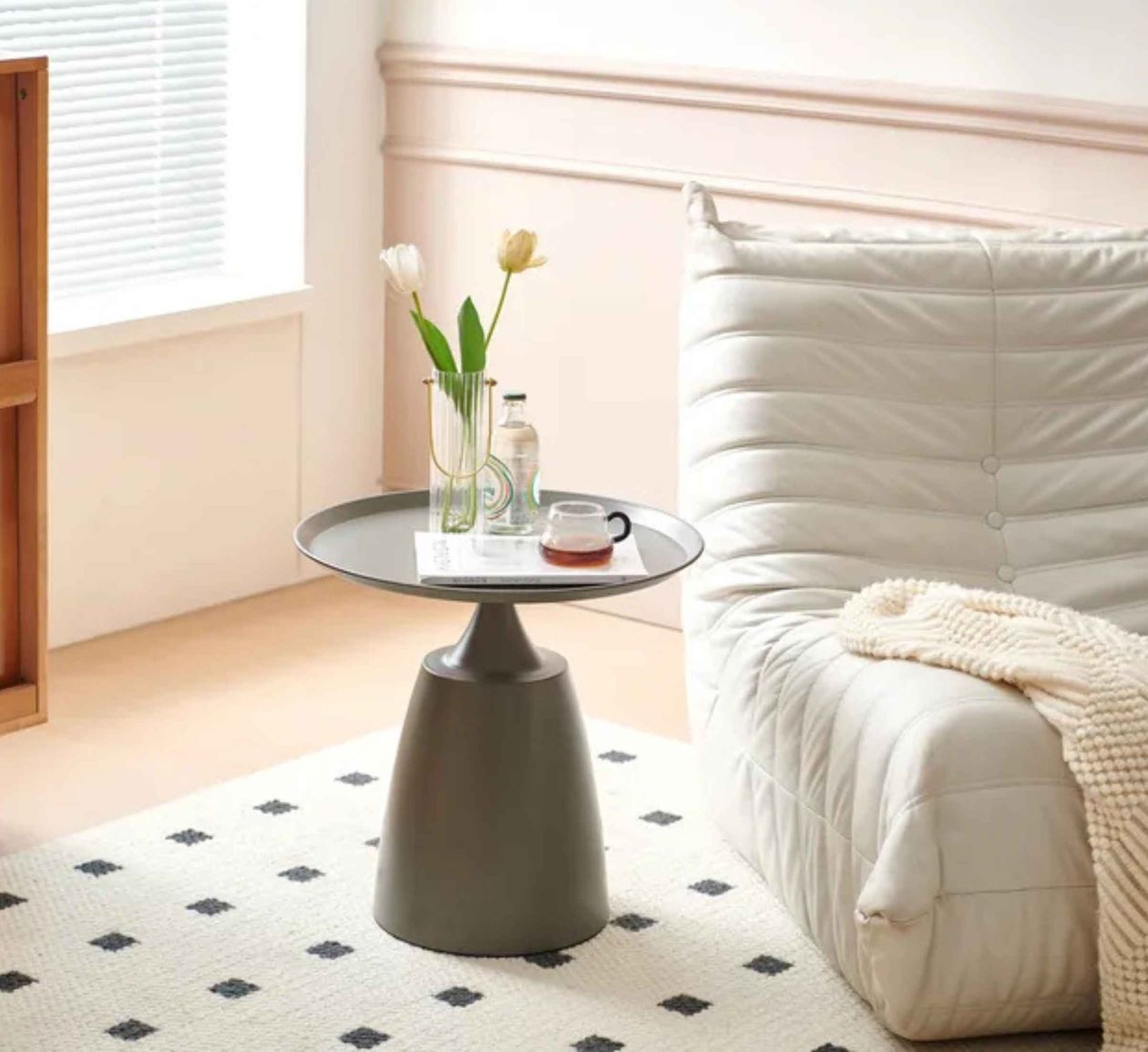 Beiger Sessel mit Decke neben einem runden, grauen Tisch mit Vase, Wasserflasche und Teetasse, auf einem cremefarbenen Teppich mit schwarzen Rauten.