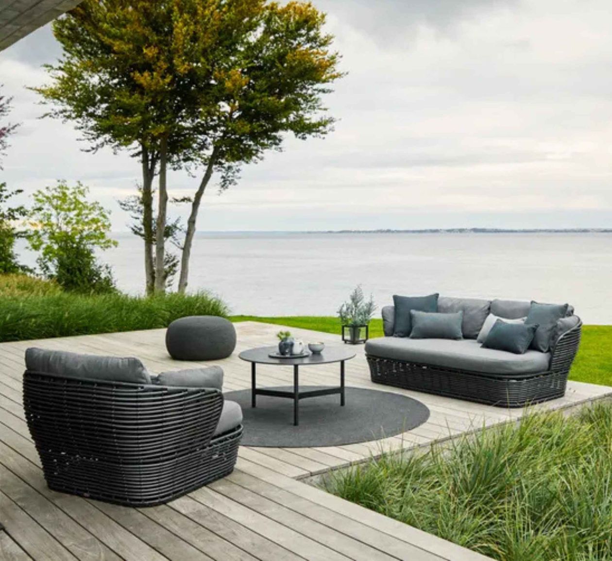 Moderne Terrasse mit grauen Outdoor-Sofas und Tisch auf Holzboden, umgeben von Rasen, Bäumen und Seeblick im Hintergrund.