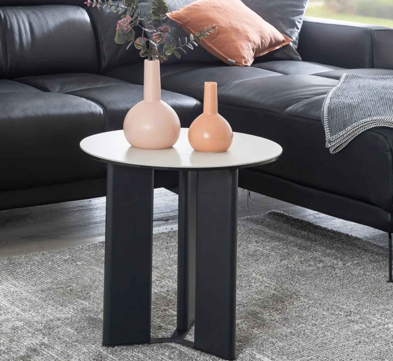 Runder Beistelltisch aus schwarzem Metall mit heller Tischplatte, zwei rosa Vasen darauf, neben einem schwarzen Ledersofa und grauem Teppich.