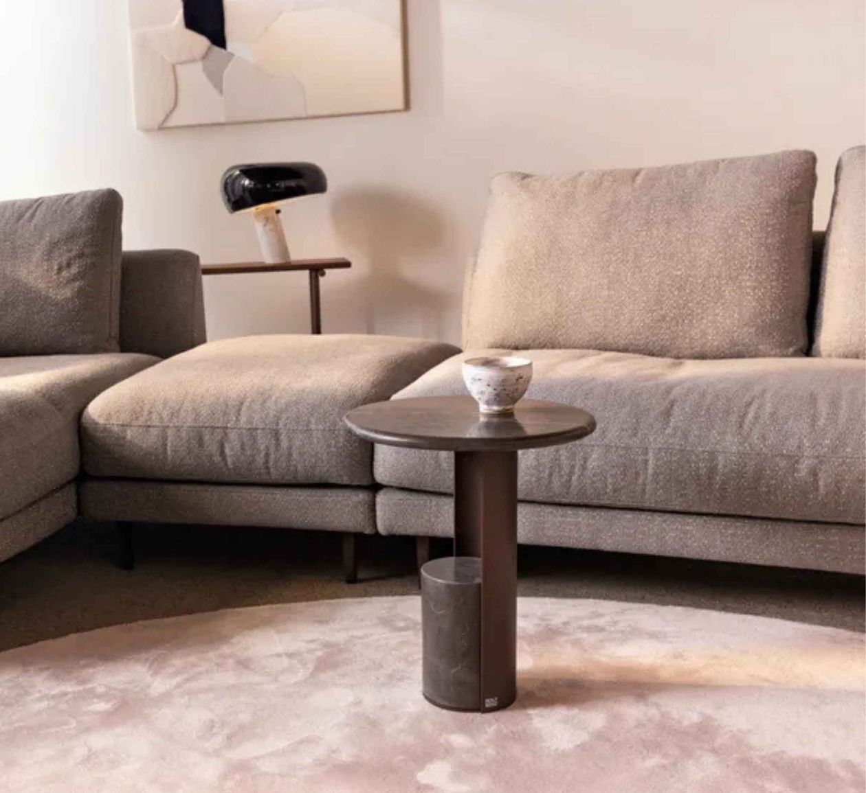Gemütliches Wohnzimmer mit grauem Sofa, kleinem runden Tisch, dekorativer Schale und modernem Kunstwerk an der Wand.