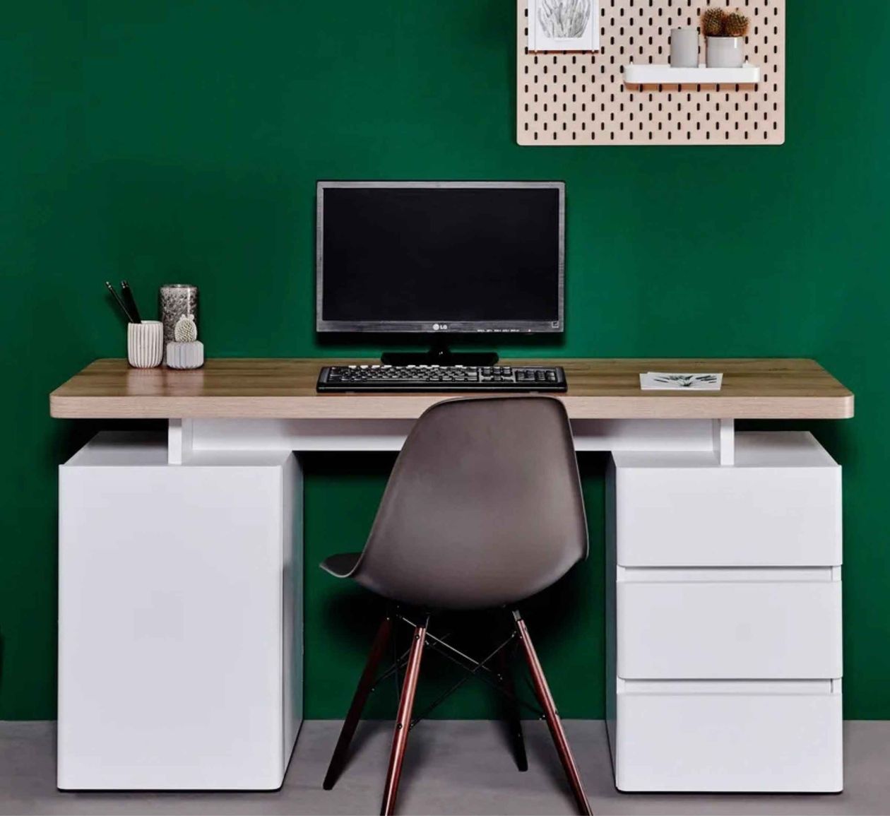 Schreibtisch mit Holzplatte, schwarzer Stuhl, Bildschirm, Tastatur, Pflanzen und Regal an grüner Wand.