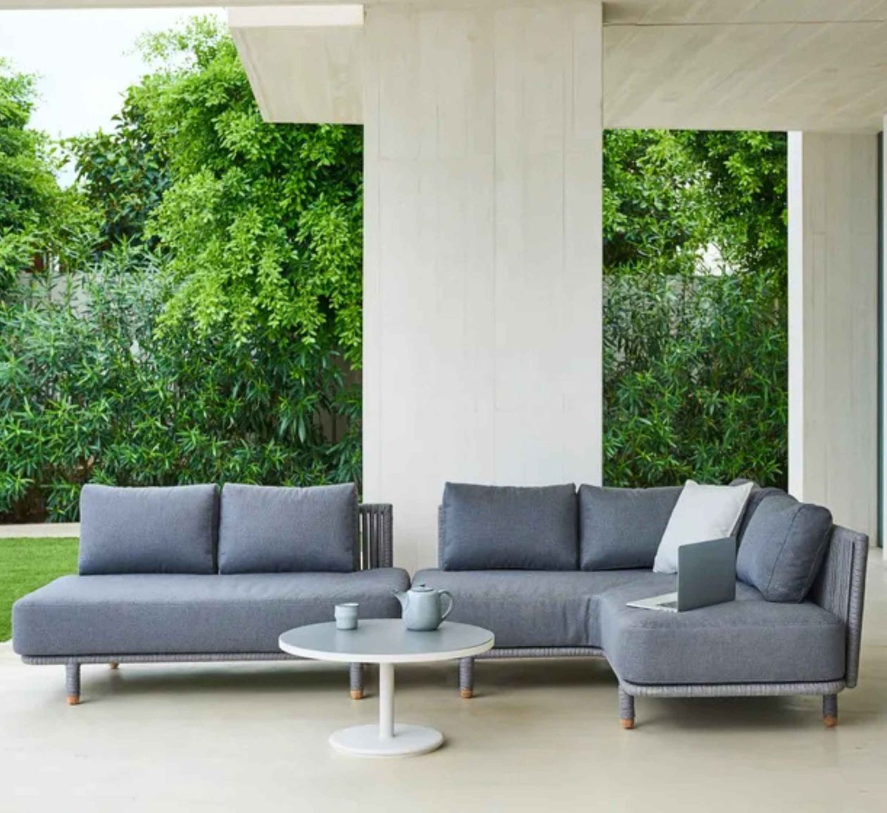 Graues Outdoor-Sofa-Set mit L-förmiger Couch, kleinem runden Tisch mit Teekanne, Tasse und Laptop, umgeben von üppigem grünen Pflanzen.