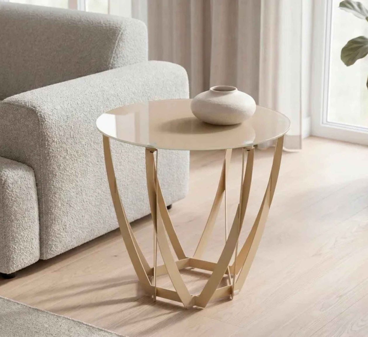 Moderner Beistelltisch mit goldenem, geometrischem Metallgestell, runder Glasplatte und dekorativer Vase, neben einem beigen Sofa vor einer Fensterfront.