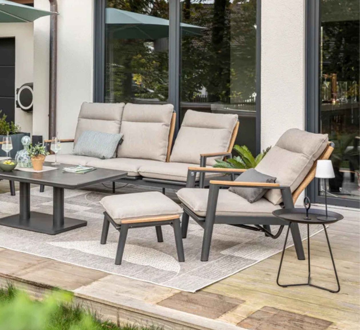 Moderne Terrassenmöbel auf einer Holzterrasse, mit beigen Kissen, einem Tisch, Pflanzen und Gläsern, vor einem Fenster mit grünem Hintergrund.