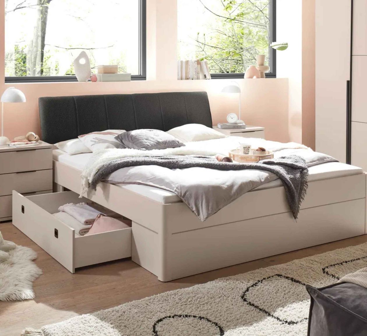 Helles Schlafzimmer mit einem Bett, das über einen Stauraum verfügt. Ein Teppich liegt auf dem Holzboden, Fenster mit Blick ins Freie.