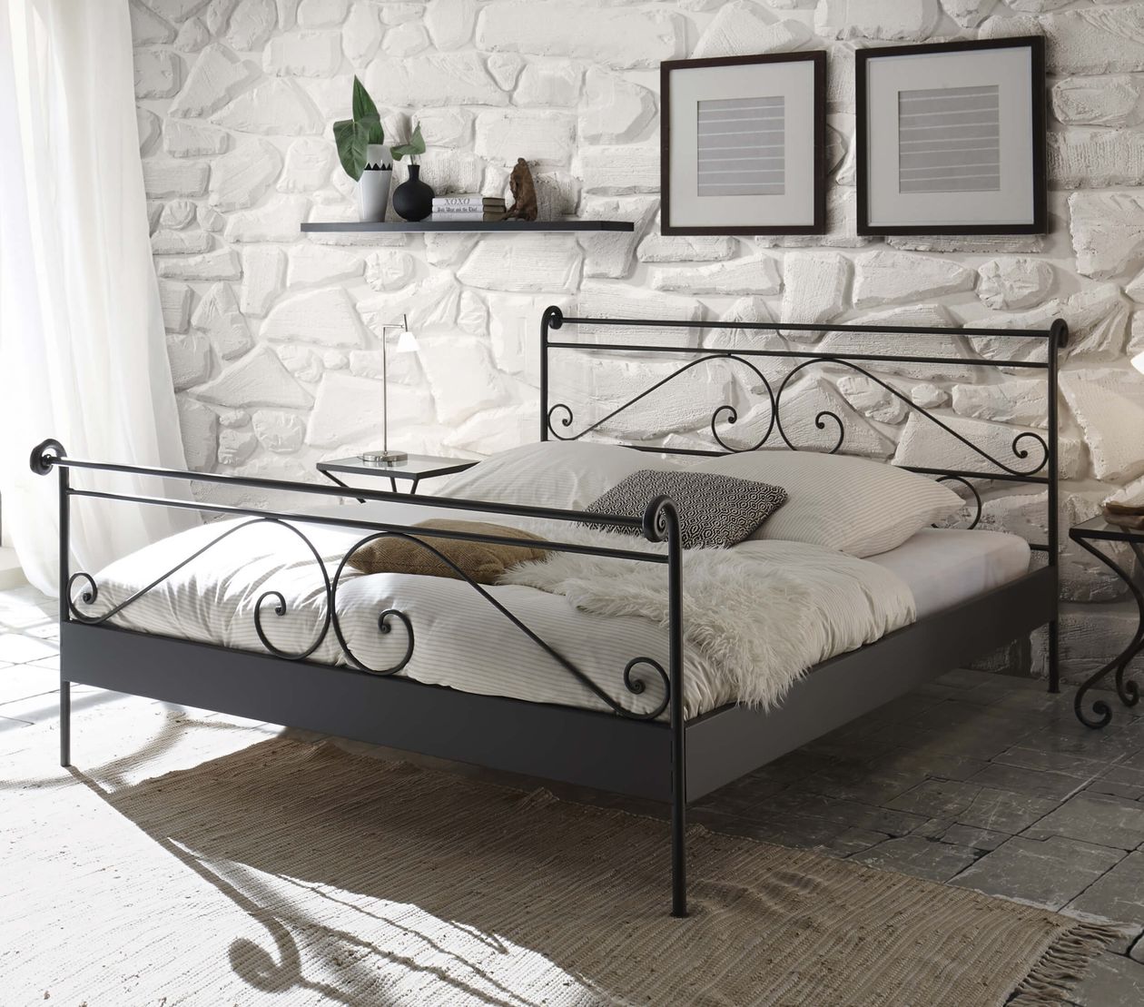 Schlafzimmer mit einem schmiedeeisernen Bett, weißen Bettwaren, Steinwand, zwei gerahmten Bildern und einem Regal mit Dekoration.