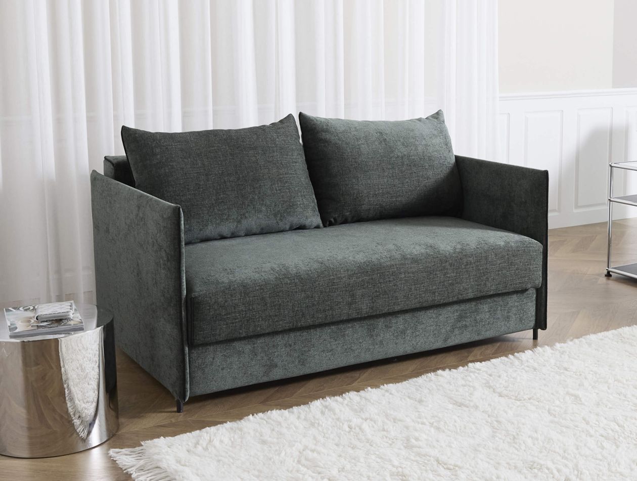 Graues Zweisitzer-Sofa mit Kissen, neben einem glänzenden Beistelltisch. Holzboden und weißer Teppich unter weißem Vorhang.