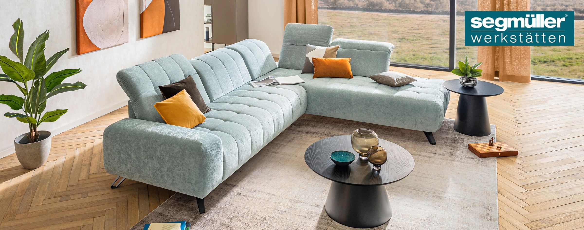 Modernes Wohnzimmer mit hellgrauem Ecksofa, bunten Kissen und Couchtisch. Ein großes Fenster bietet Blick auf die Landschaft. Pflanzen und Kunstwerke ergänzen den Raum.