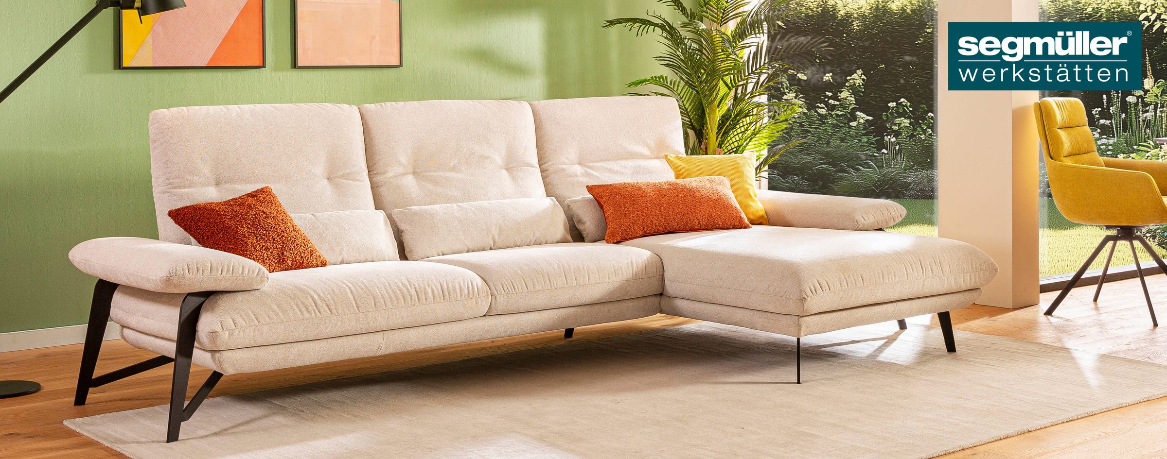 Ecksofa Denise aus den SEGMÜLLER WERKSTÄTTEN mit cremefarbenem Stoffbezug und schwarzen Füßen vor einer grünen Wand.