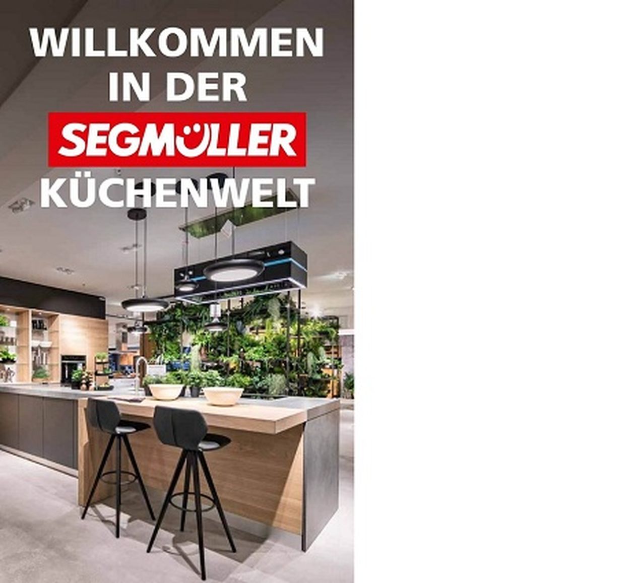 Moderner Küchenausstellungsraum mit Kochinsel, Barhockern und Grünpflanzen. Text: „Willkommen in der Segmüller Küchenwelt.“
