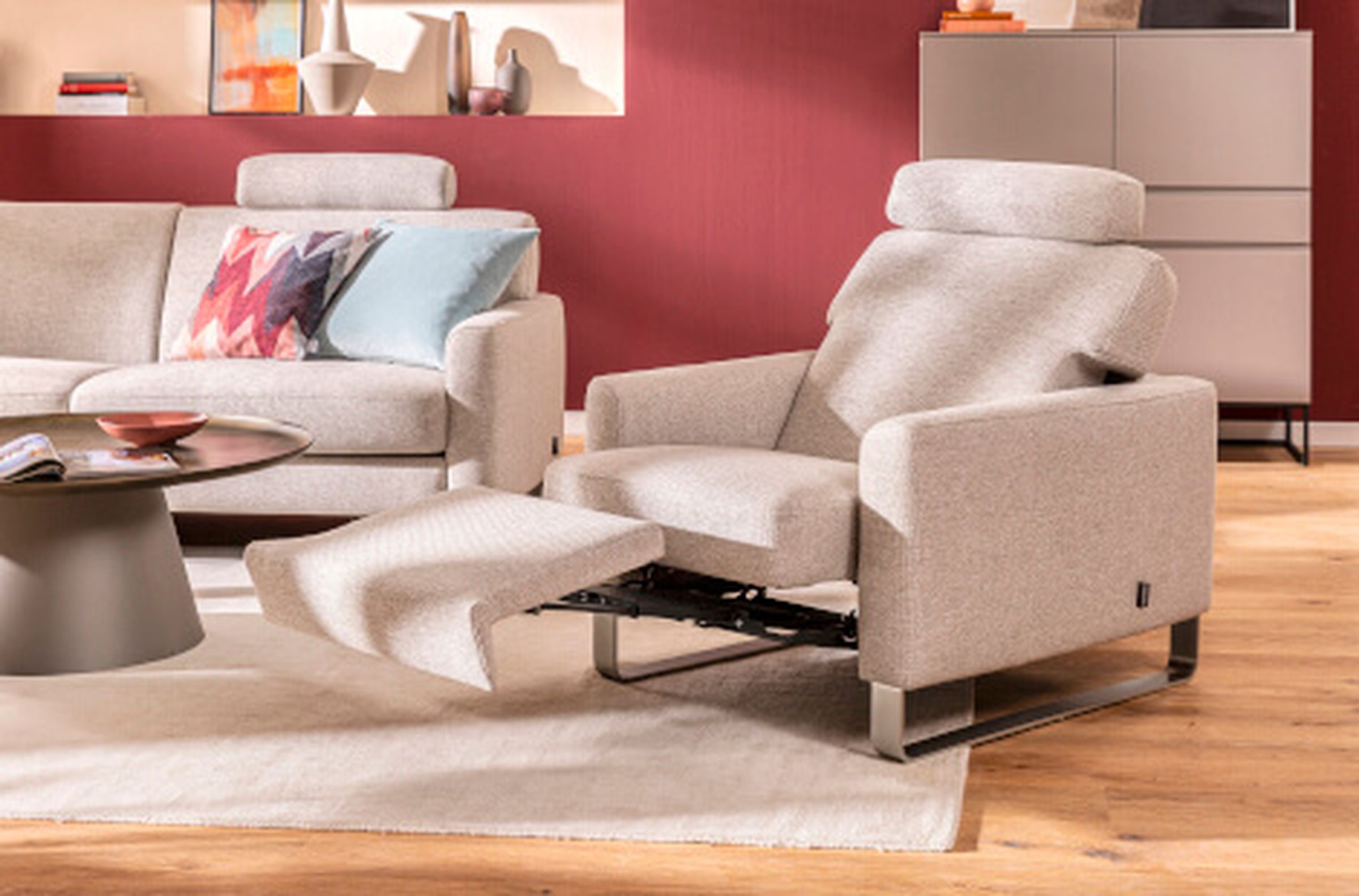 Modernes Wohnzimmer mit hellgrauem Ruhesessel, passendem Sofa, bunten Kissen, rundem Couchtisch und einer roten Akzentwand.