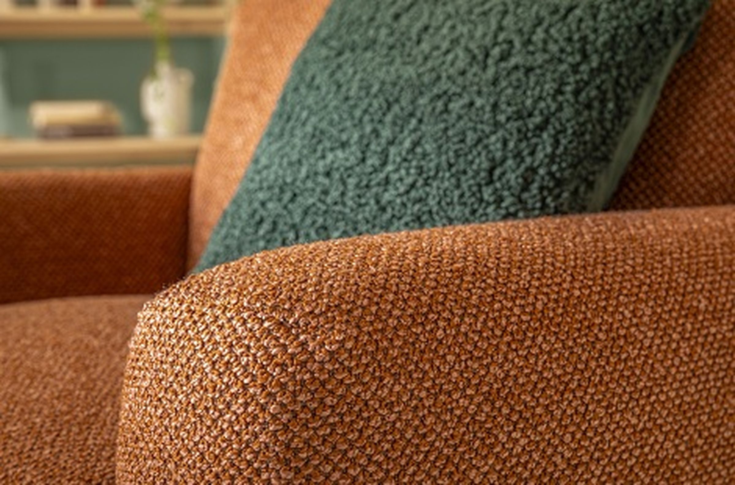 Nahaufnahme eines strukturierten orangefarbenen Sessels mit einem weichen, dunkelgrünen Kissen. Im verschwommenen Hintergrund sind ein Regal mit einem Buch und einer Vase zu sehen.