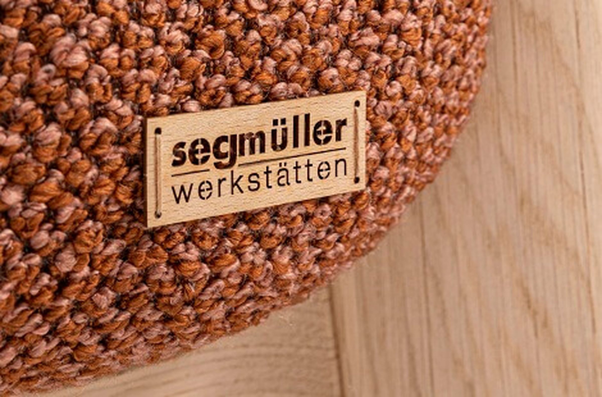 Nahaufnahme eines strukturierten braunen Strickstoffs mit einem daran befestigten Holzschild mit der Aufschrift „Segmüller Werkstätten“.