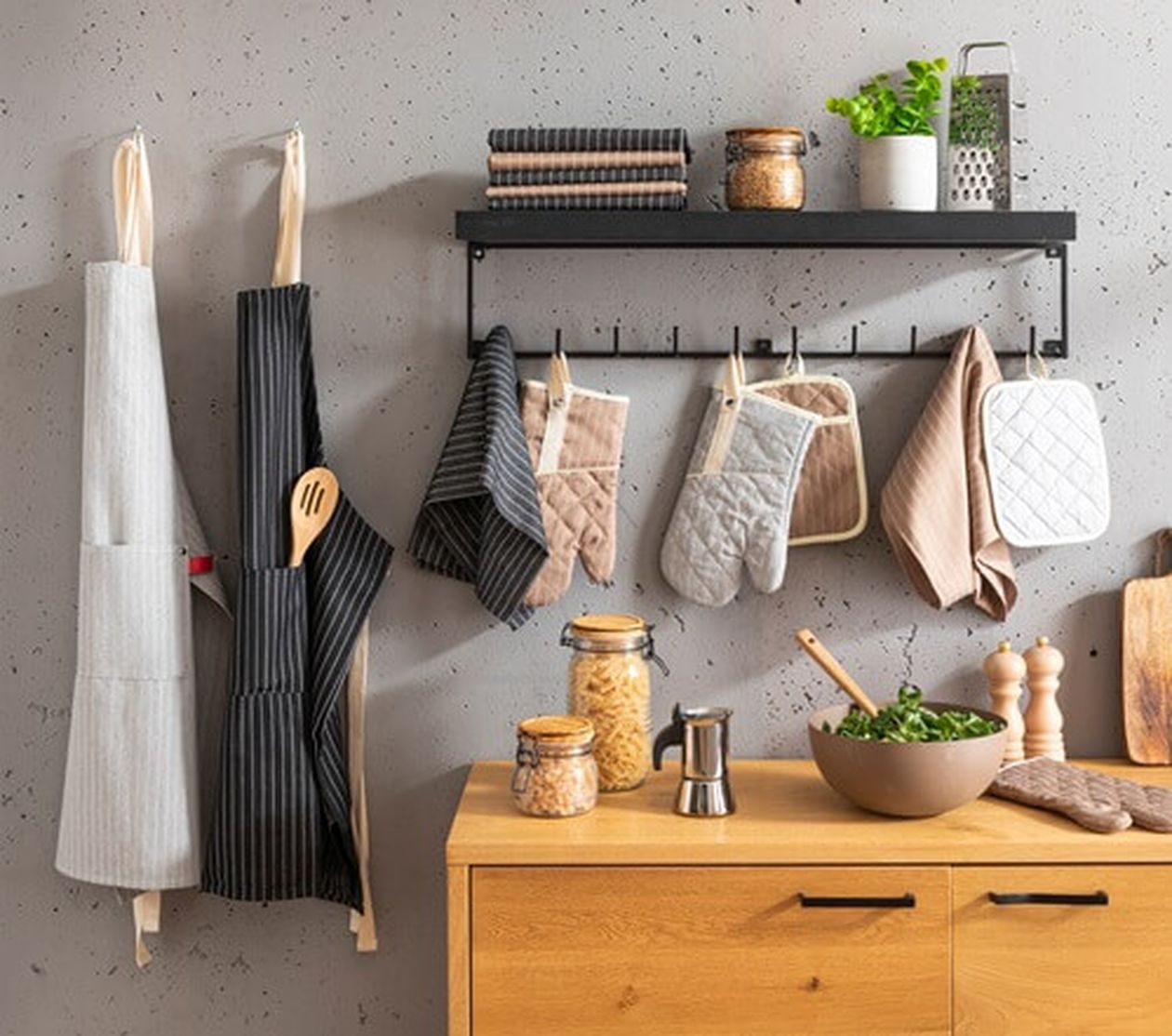 Verschiedene Küchenaccessoires wie Schürzen, Ofenhandschuhe und Topflappen hängen ordentlich an einer Leiste an der Wand über einer Holz-Arbeitsfläche. Im Vordergrund stehen Schalen, Gläser und Gewürze.