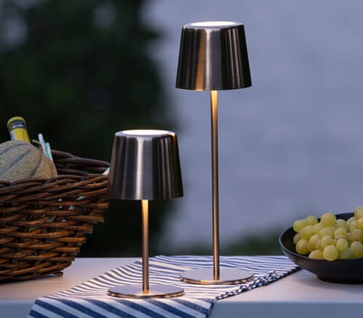 Zwei elegante, metallische Tischlampen auf einem gestreiften Tuch neben einem Korb mit einer Melone und einer Schale Weintrauben im Freien.