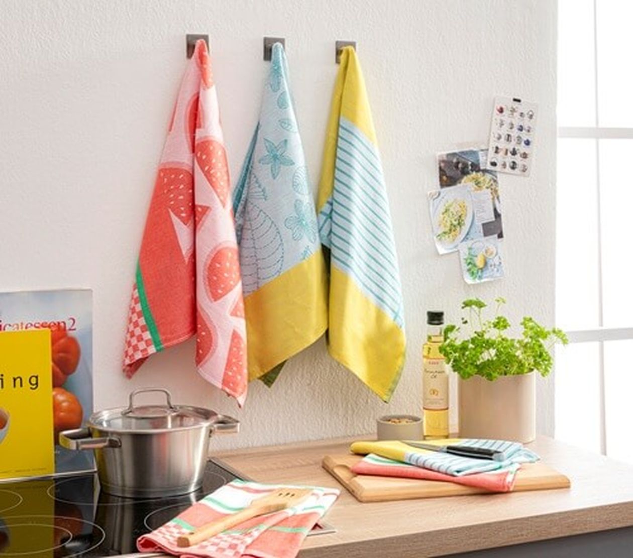 Mehrfarbige Geschirrtücher hängen nebeneinander an Haken in einer hellen Küche. Auf der Arbeitsfläche liegen weitere Tücher und Küchenutensilien, dazu ein Topf und frische Kräuter.