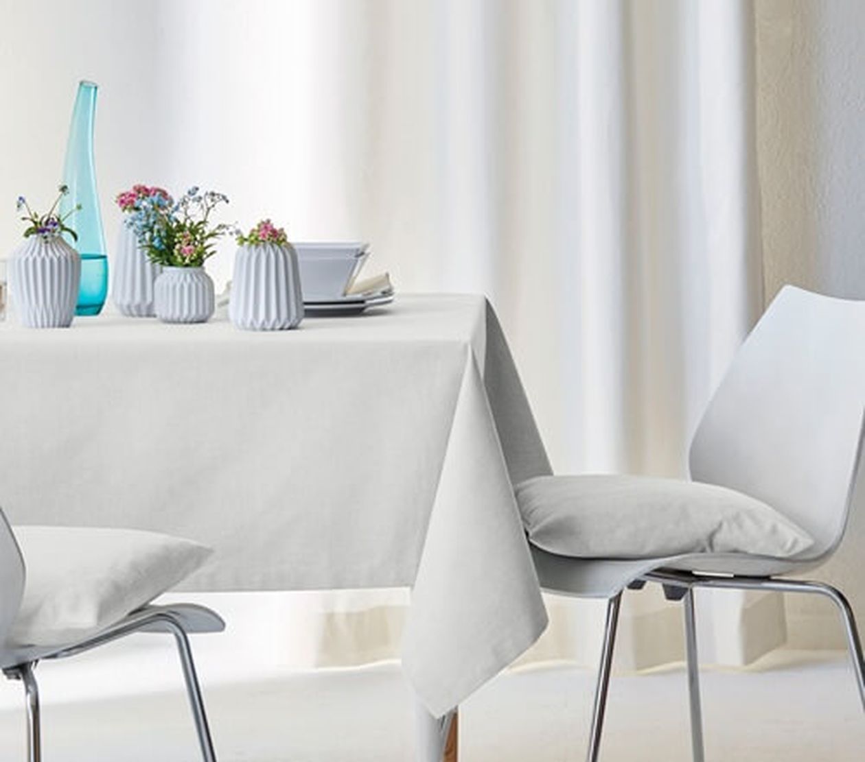 Helle, schlichte Tischdecke auf einem Tisch, der mit dezentem weißem Geschirr und kleinen Blumenvasen dekoriert ist. Neben dem Tisch steht ein weißer Stuhl mit Kissen.