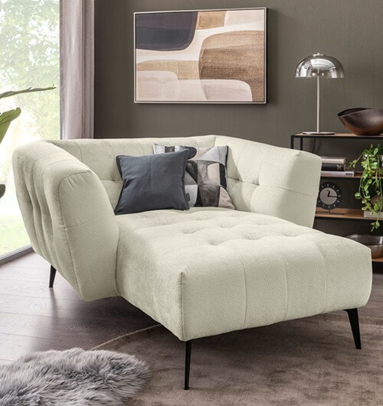 Modernes Wohnzimmer mit cremefarbener Chaiselongue, abstrakter Wandkunst, Metalllampe und einer Pflanze auf einem Regal. Licht fällt durch ein Fenster.