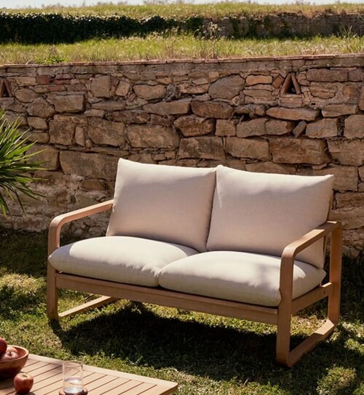 Ein beigefarbenes, gepolstertes Outdoor-Sofa mit Holzrahmen steht auf Gras, dahinter eine Steinmauer. Im Vordergrund ist teilweise ein Holztisch sichtbar.