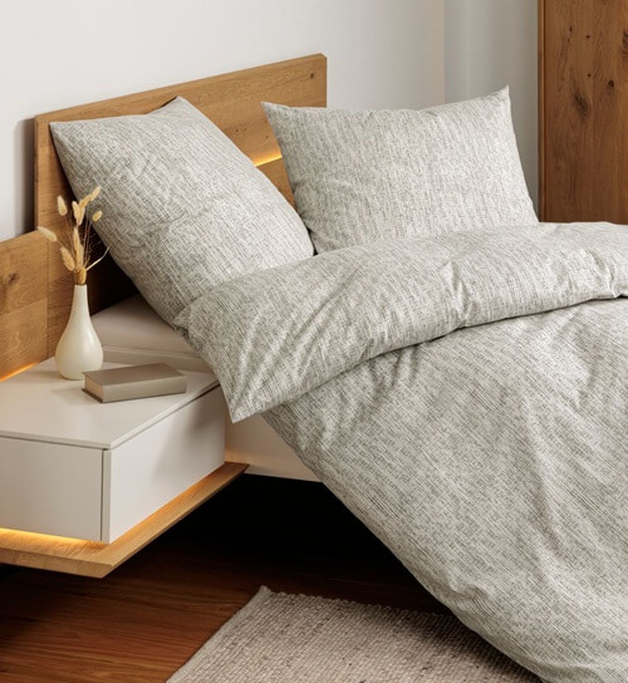 Modernes Schlafzimmer mit Holzkopfteil, grauer Strukturbettwäsche und einem schwebenden Nachttisch mit Vase und Büchern. Sanfte Beleuchtung verstärkt die gemütliche Atmosphäre.