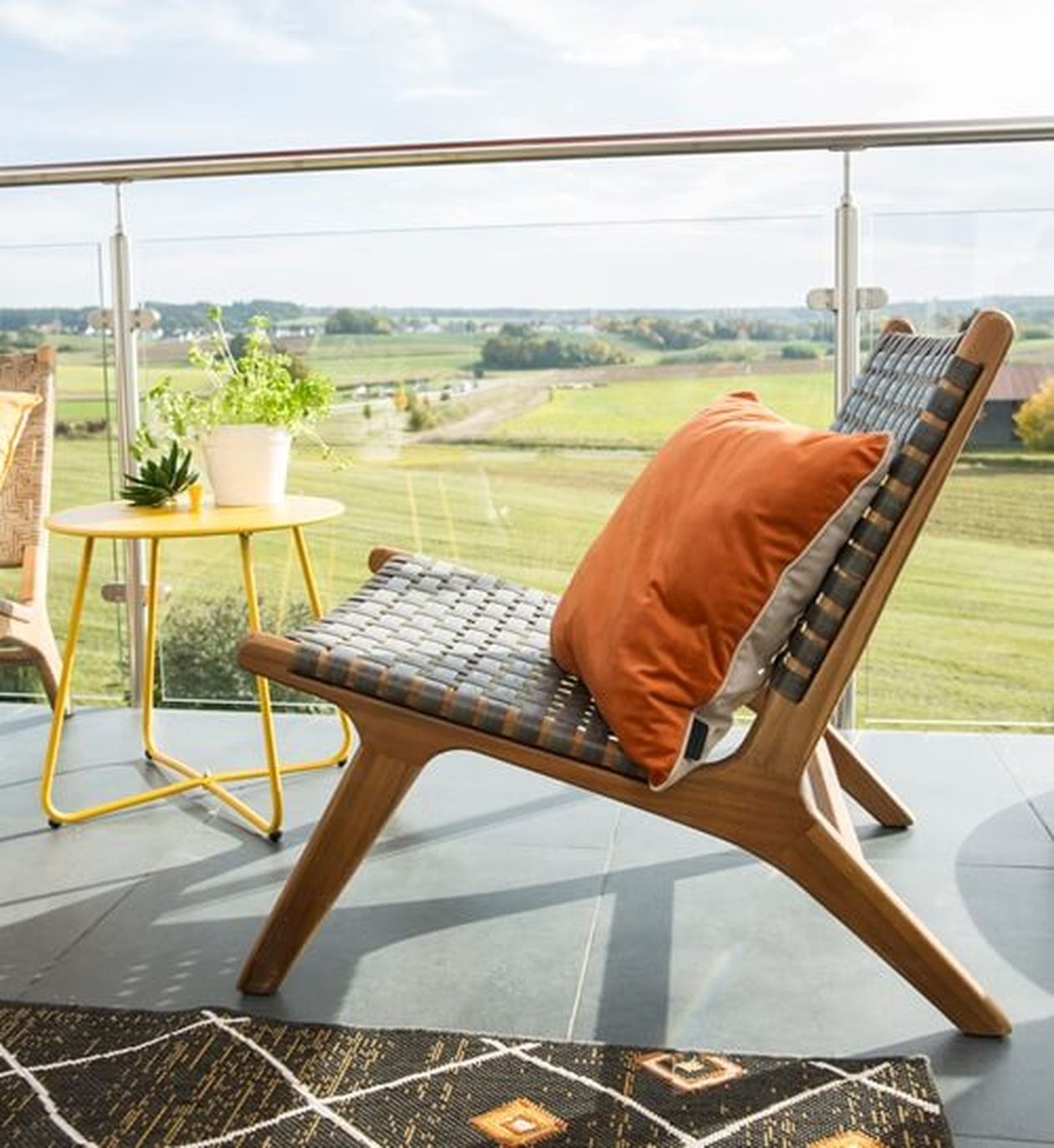 Ein Holzstuhl mit orangefarbenen und grauen Kissen auf einem Balkon, neben einem gelben Tisch mit Topfpflanzen und Blick auf eine grüne Landschaft.