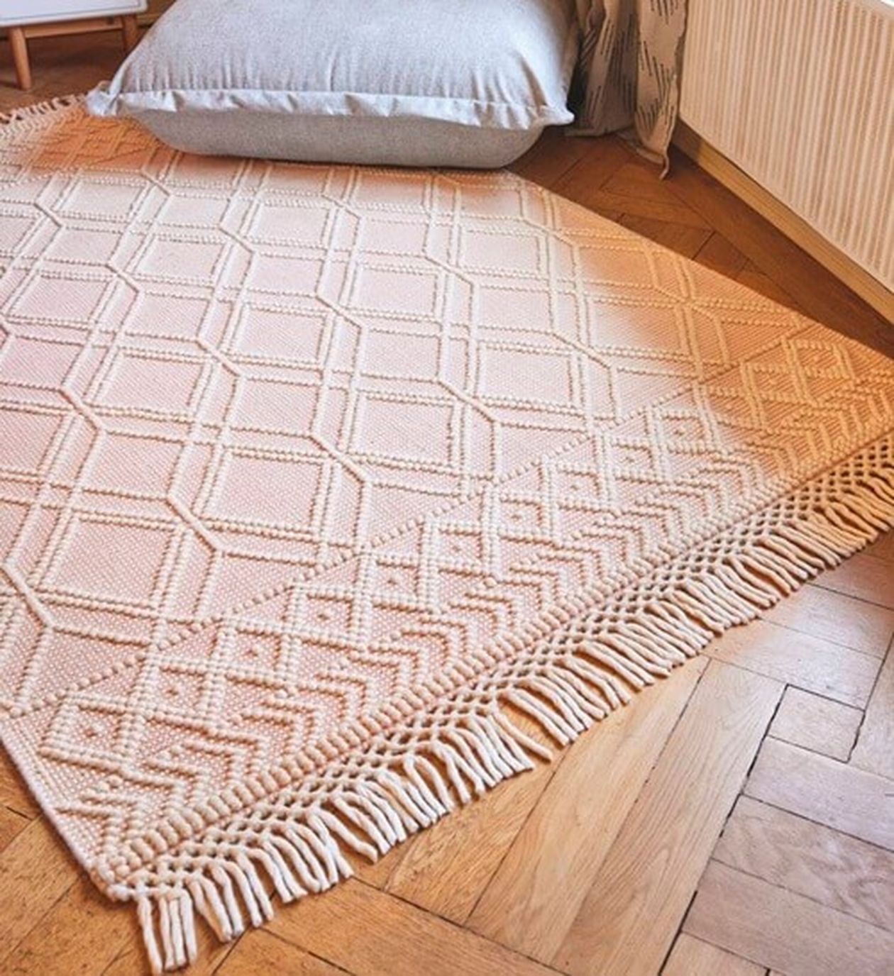 Ein beigefarbener gewebter Teppich mit geometrischen Mustern und Fransenkanten auf einem Holzboden, neben einem großen grauen Kissen.