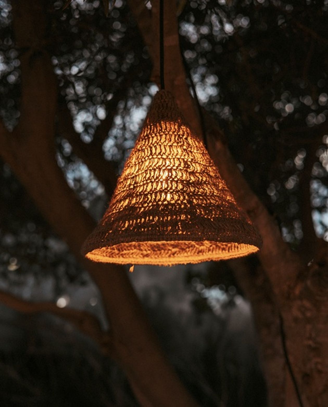 Eine geflochtene, kegelförmige Lampe strahlt ein warmes Licht aus und hängt in einer schwach beleuchteten Außenumgebung an einem Ast.