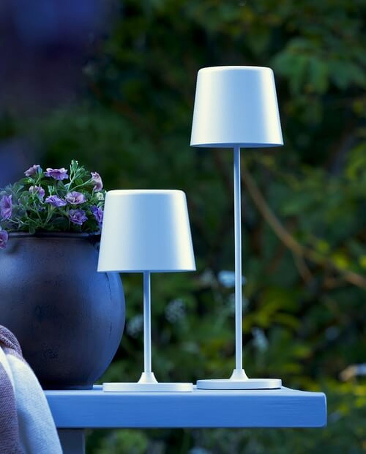 Zwei weiße Tischlampen unterschiedlicher Höhe auf einer blauen Fläche, neben einem Topf mit lila Blumen, mit einem verschwommenen grünen Hintergrund.