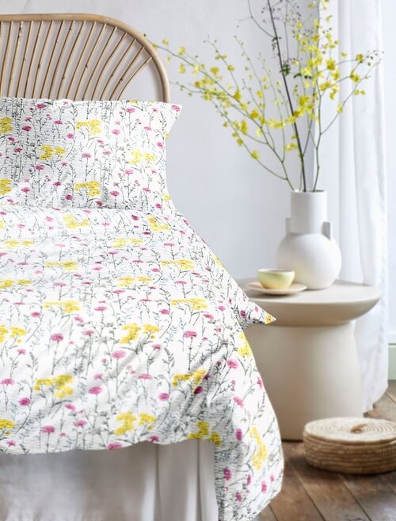 Bettwäsche mit Blumenmuster und gelben und rosa Blumen auf einem Bett, daneben ein runder Tisch mit einer Vase mit gelben Blumen und einem geflochtenen Korb.