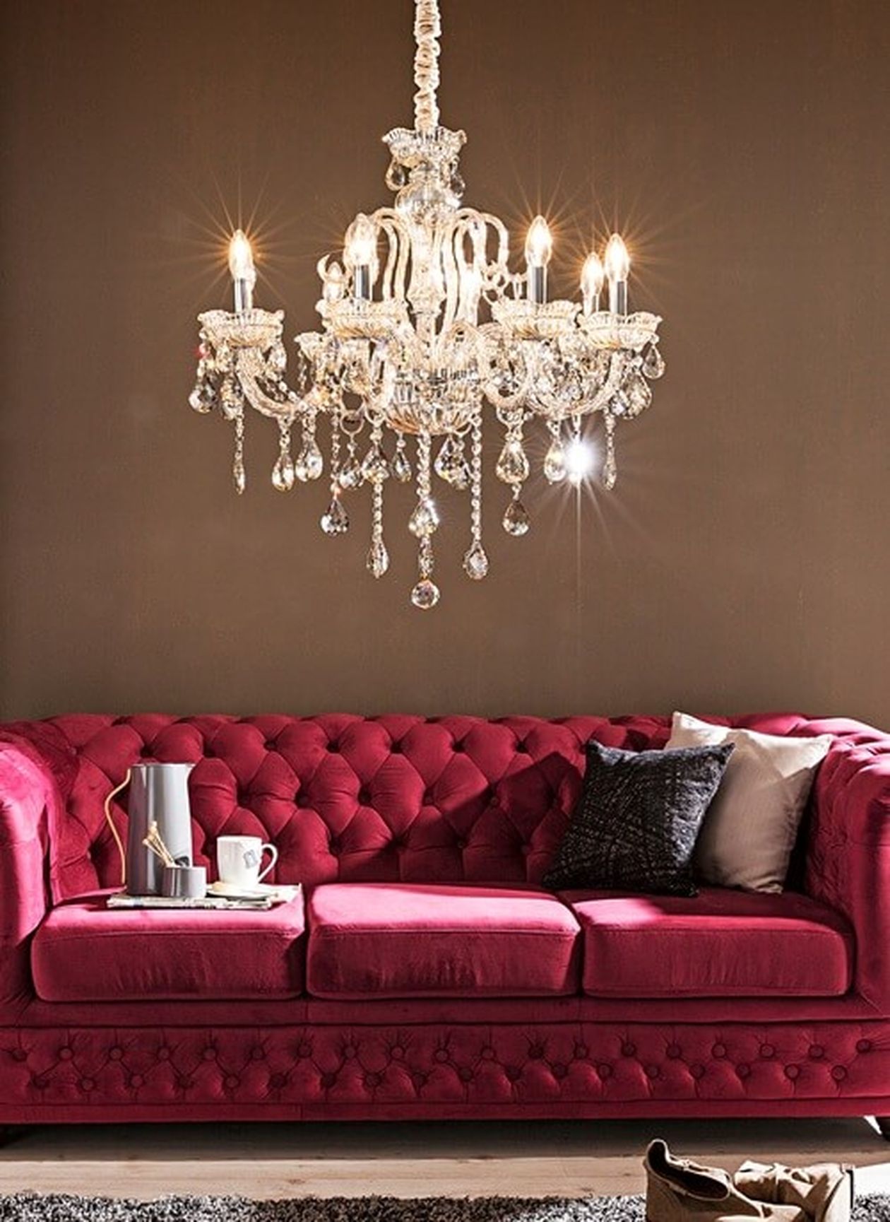 Eleganter Kristallleuchter über einem weichen, rosafarbenen Sofa mit Polstern vor einer braunen Wand. Auf dem Sofa steht ein Kaffeeservice.