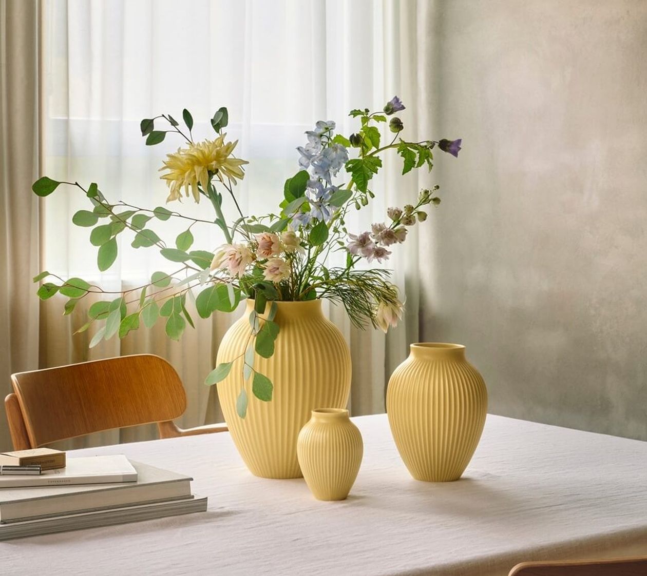 Drei gelbe, geriffelte Keramikvasen auf einem gedeckten Tisch vor einem Fenster mit hellen Vorhängen. In einer Vase steht ein bunter Frühlingsstrauß.