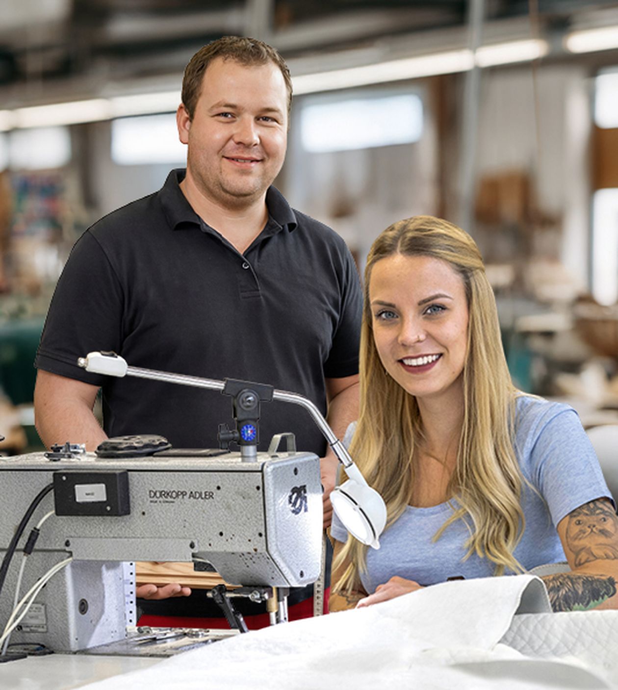 Ein älterer Mann und eine junge Frau arbeiten gemeinsam an einer industriellen Nähmaschine in einer Werkstatt.