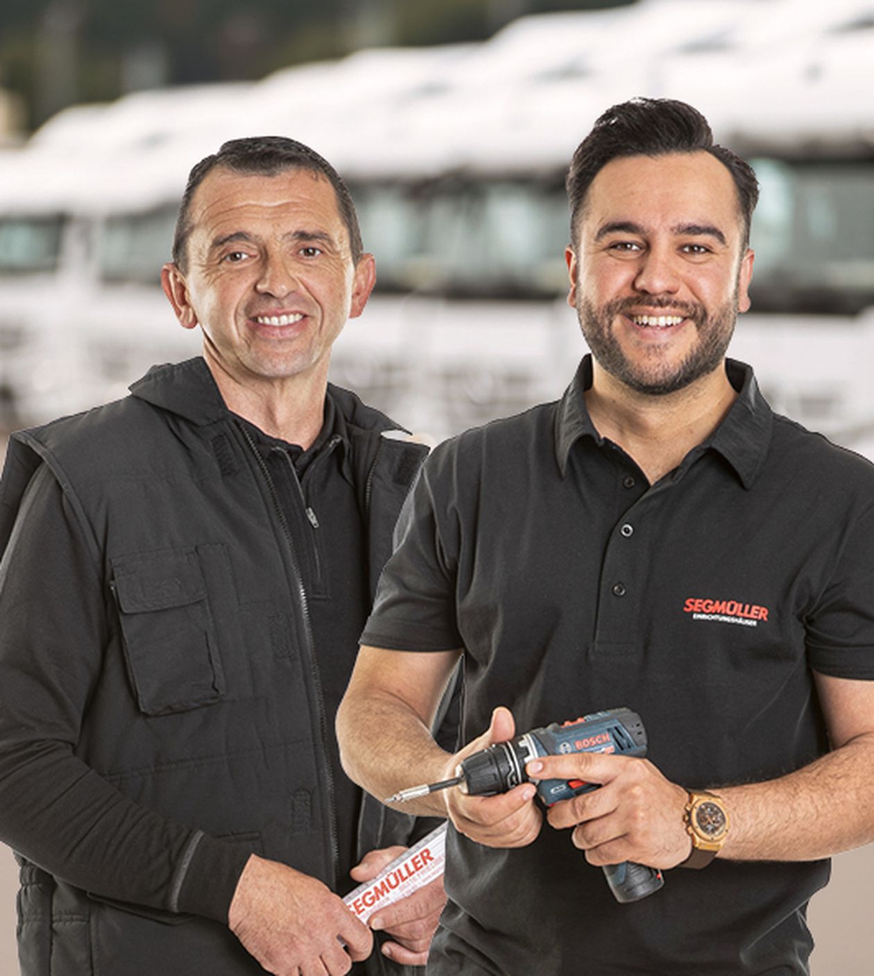 Zwei Mechaniker in Arbeitskleidung halten Werkzeuge und stehen lächelnd in einem Logistikzentrum.