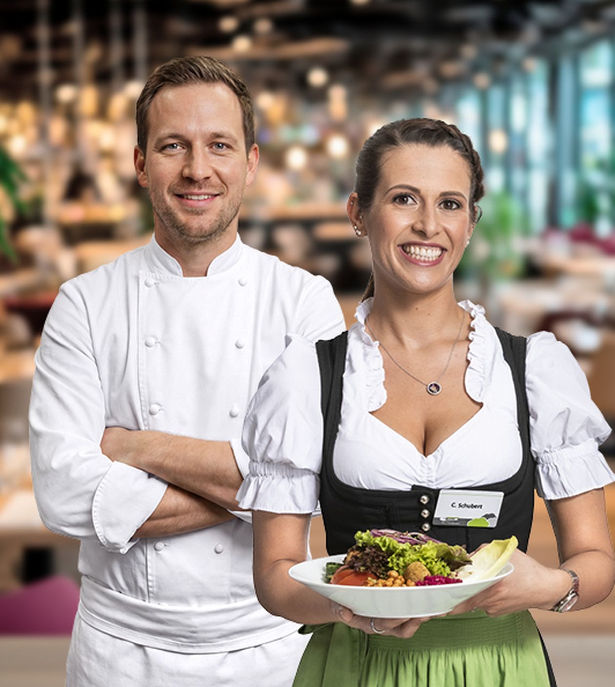 Ein Koch und eine Servicekraft in traditioneller Tracht präsentieren lächelnd ein Teller mit frischem Salat in einem Restaurant.