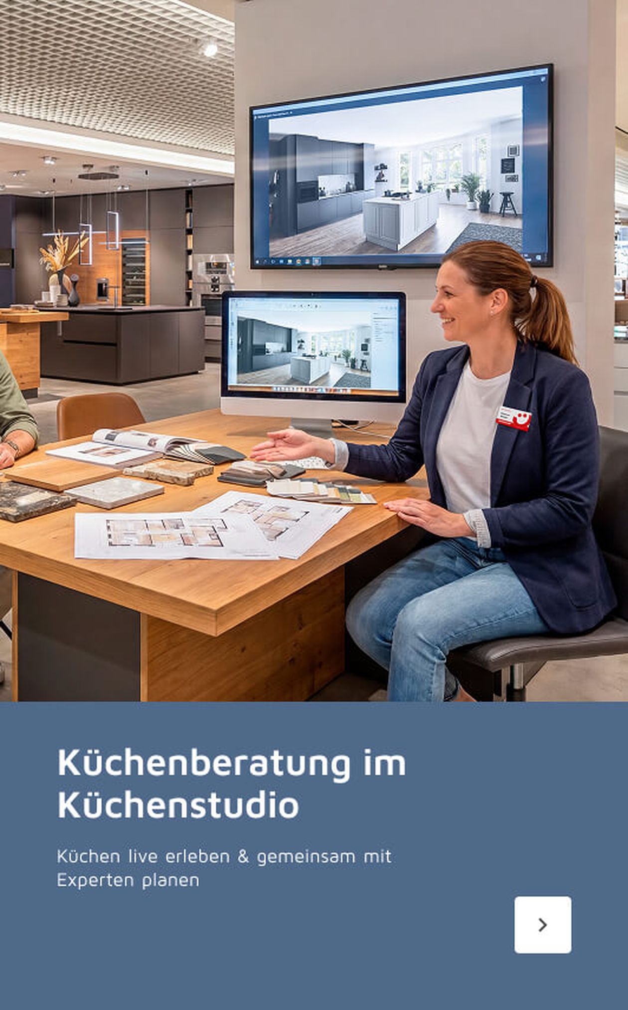 Beraterin im Küchenstudio sitzt an einem Tisch mit Plänen und Mustern, während Bilder von Küchen auf Bildschirmen im Hintergrund zu sehen sind.