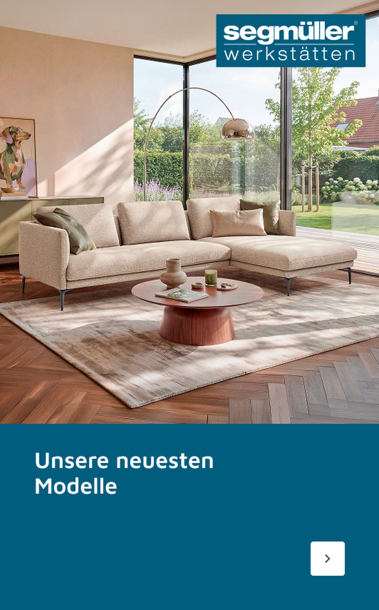 Modernes Wohnzimmer mit beigem Sofa, Holztisch, großer Fensterfront und Gartenblick. Segmüller-Werbung: "Unsere neuesten Modelle".