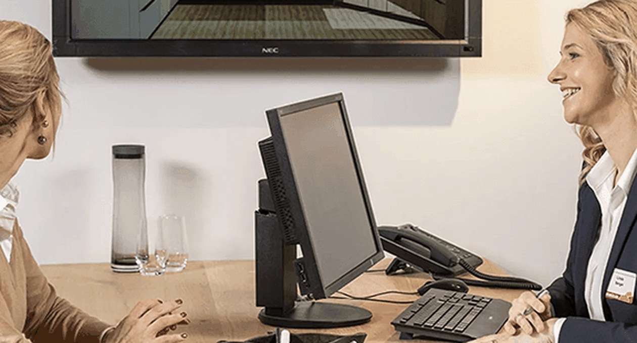 Zwei Frauen in einem Büro, betrachten einen Computermonitor. Im Hintergrund ist ein Bildschirm mit einer Küchenplanung sichtbar.