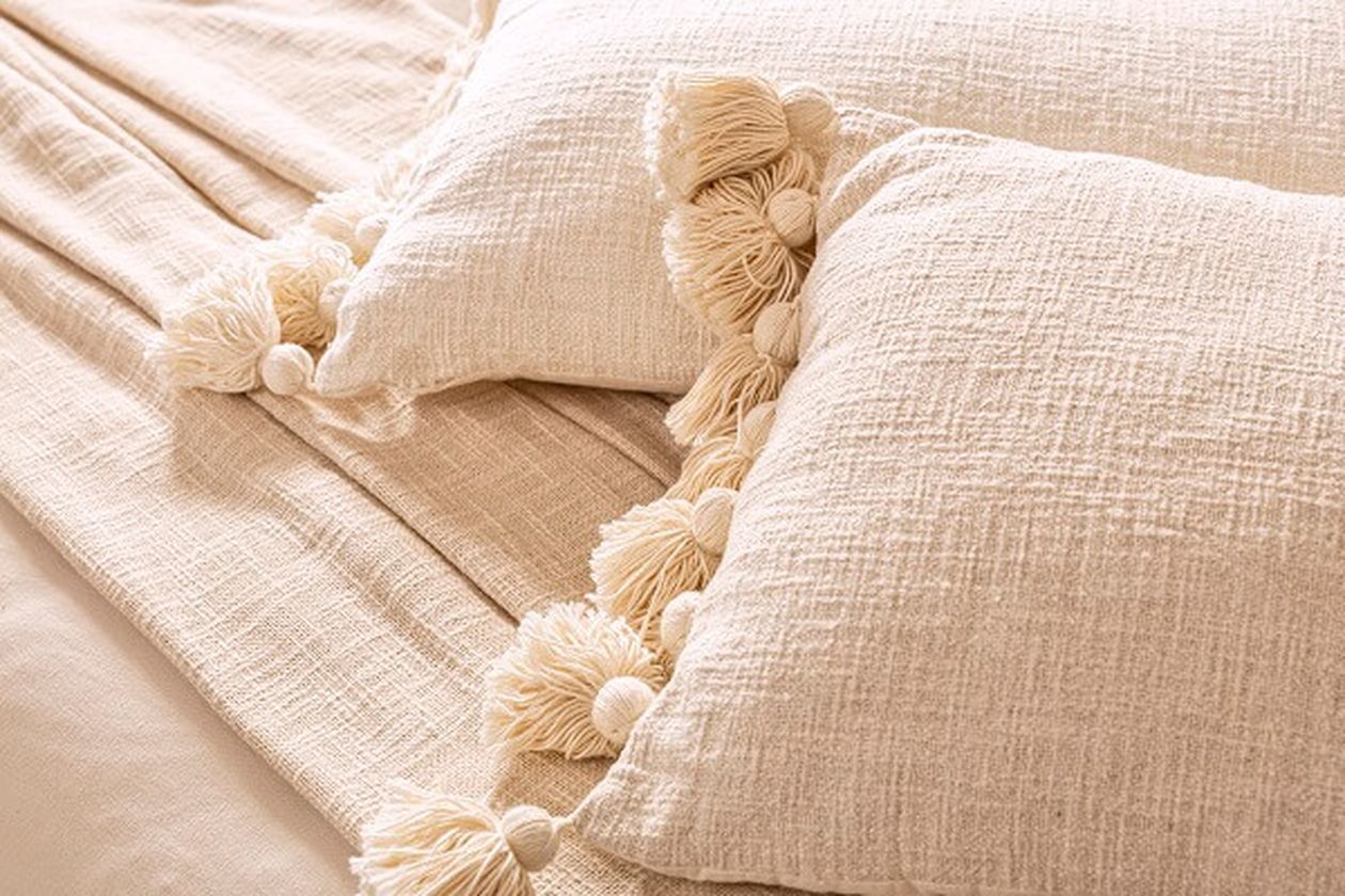 Zwei beige Dekokissen mit dekorativen Quasten liegen auf einem Bett.