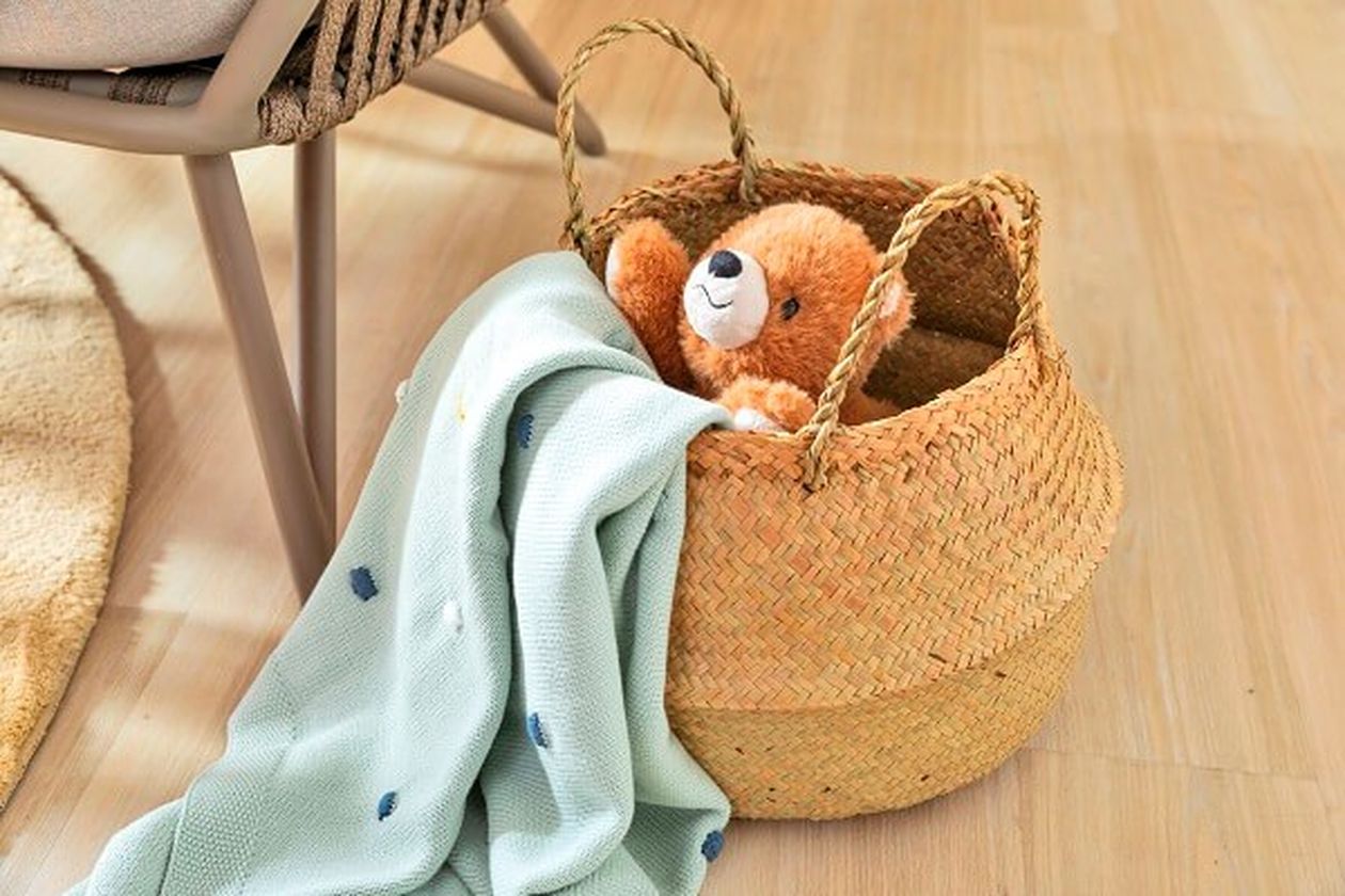 Auf dem Boden platzierter Korb aus Geflecht mit einem Teddybären und einer hellblauen Decke darin.