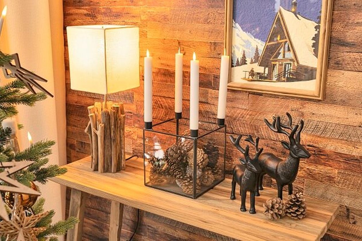 Gemütlicher Holztisch mit brennenden Kerzen, einer Lampe, Tannenzapfen, Rentierfiguren und einem Bild einer verschneiten Hütte, der eine warme, festliche Atmosphäre schafft.