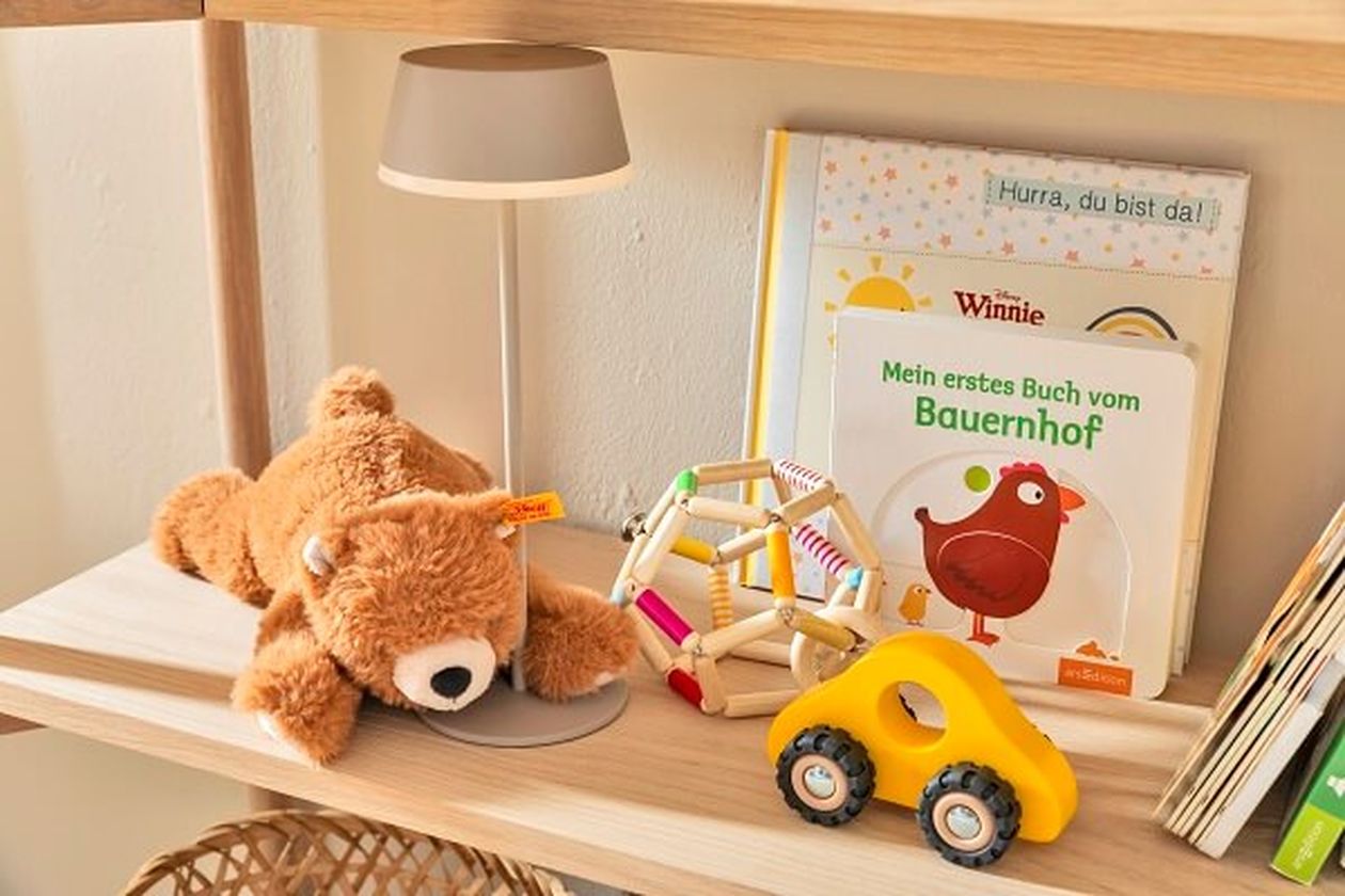 Bücherregal aus hellem Holz mit Babyzimmer-Deko: einem Teddybär, einer Tischleuchte, Spielzeug und Büchern.
