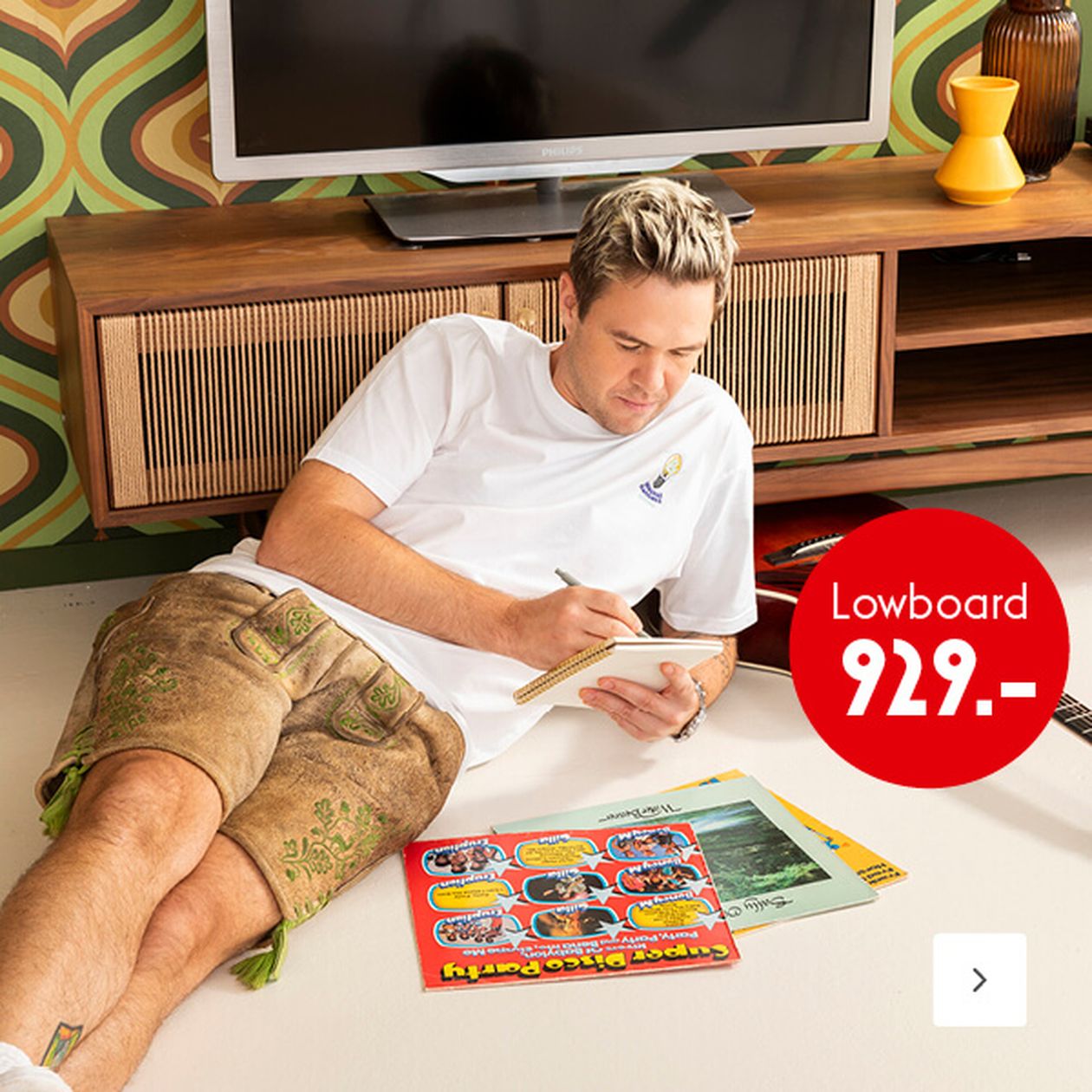 Ein Mann in weißem Hemd und Shorts sitzt auf dem Boden und schreibt in ein Notizbuch, neben ihm liegen Schallplatten-Cover. Auf dem Preisschild steht „Lowboard 929.-“. Im Hintergrund ist eine Einrichtung im Retro-Stil zu sehen.