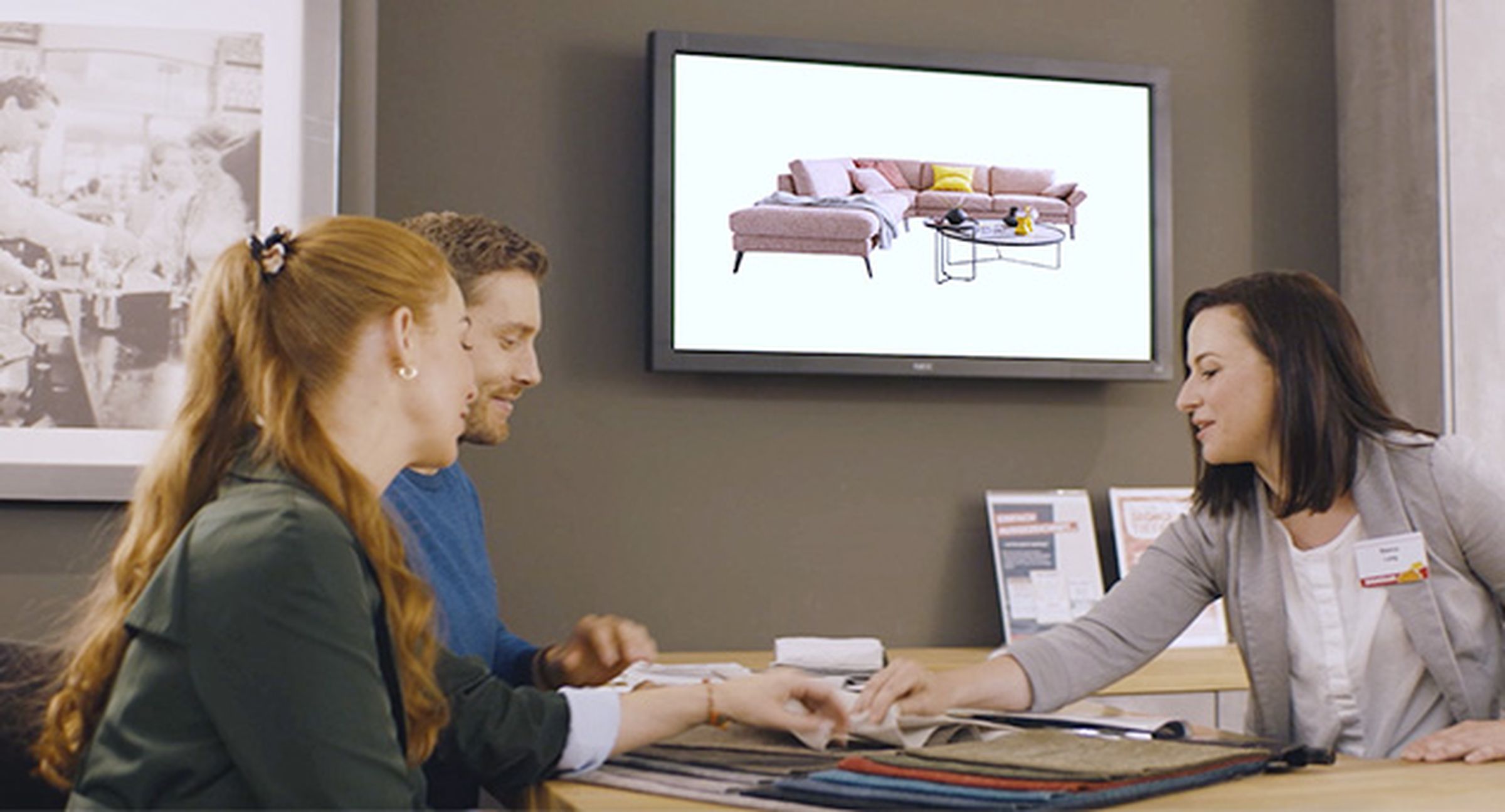 Eine Beraterin zeigt einem Paar verschiedene Stoffmuster. Im Hintergrund ist das Sofa Lipari auf einem Bildschirm zu sehen.