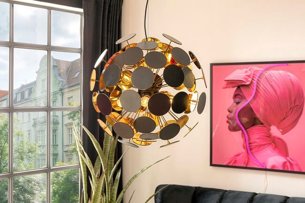 Eine moderne, kugelförmige Pendelleuchte mit goldenen Akzenten und schwarzen Scheiben hängt vor einem Fenster mit Stadtblick. In der Nähe befindet sich ein neonpinkes Wandbild.