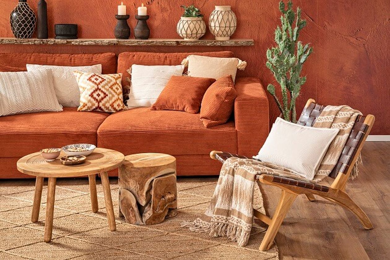 Gemütliches Wohnzimmer mit orangefarbenem Sofa, gemusterten Kissen, Holztisch und -stuhl, Kerzen und einer grünen Pflanze vor einer strukturierten Terrakottawand.