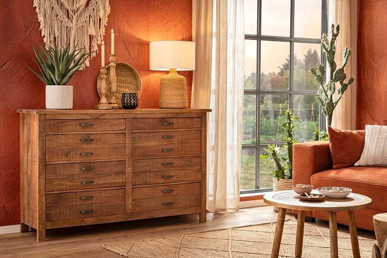 Gemütliches Wohnzimmer mit Holzkommode, Dekorationsgegenständen, orangefarbenem Sofa, rundem Couchtisch und großem Fenster mit transparenten Vorhängen und Blick ins Freie.