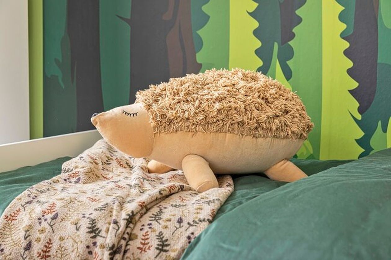 Ein Plüschigel mit geschlossenen Augen liegt auf einem grünen Bett neben einer gemusterten Decke vor einem Hintergrund mit Blättertapete.