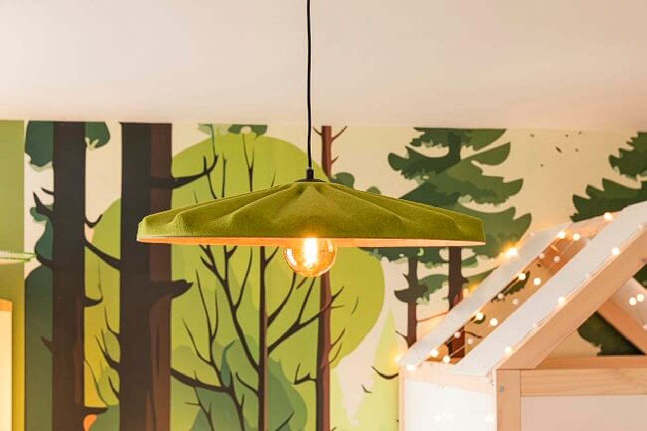 Grüne Hängelampe mit freiliegender Glühbirne vor einer Tapete mit Waldmotiv und einem festlich mit Lichterketten geschmückten Hochbett.