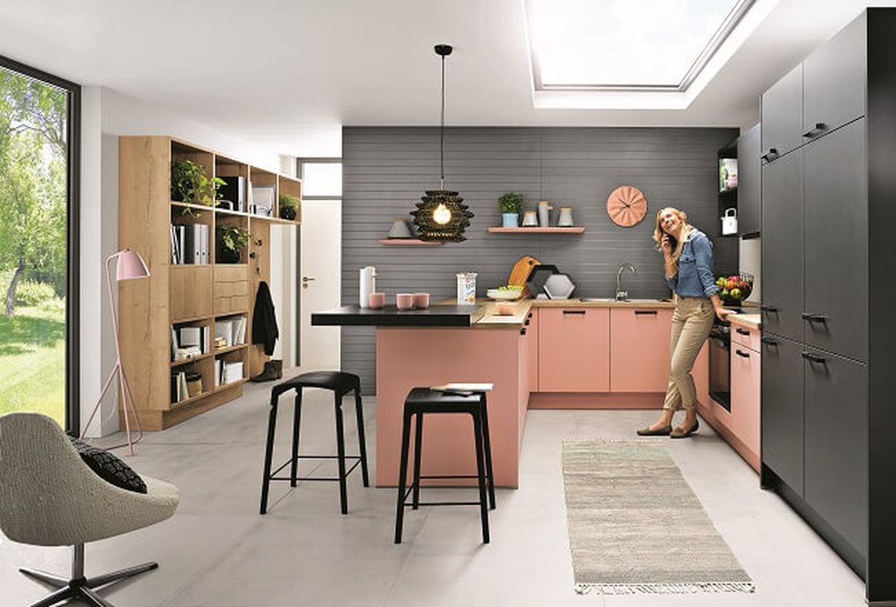 Moderne Küche in Pink und Schwarz mit einer Insel, zwei Hockern, Regalwand, Teppich und einer Person am Handy stehend. Großer Lichteinfall.