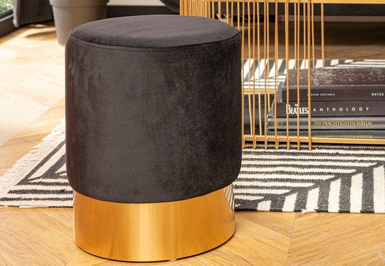 Ein schwarzer Samthocker mit goldenem Fuß auf einem Holzboden, neben einem goldenen Drahtgestelltisch und einem gemusterten Teppich.
